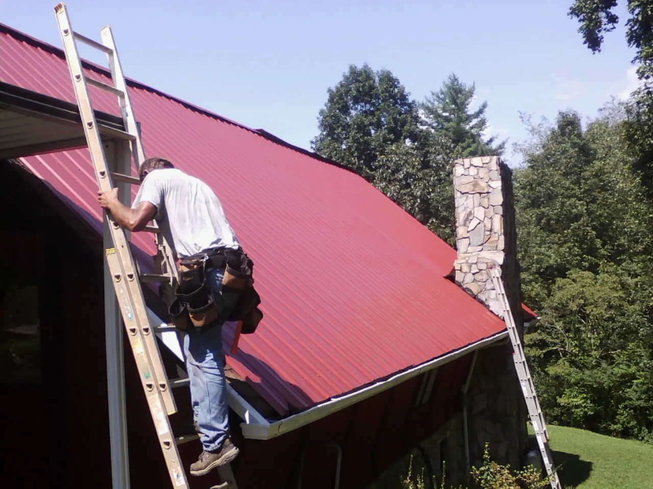Roofer on a ladder beside a red metal roof and stone chimney — Soffit & Fascia Repair by Roofing Brothers in Moorland